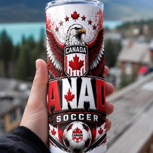 May include: A white and red insulated tumbler featuring a Canadian flag design with an eagle, maple leaves, and the words "CANADA SOCCER." The tumbler has a silver straw and is held by a person.