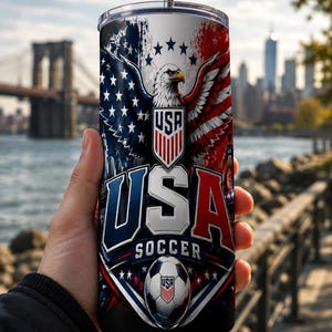 May include: A stainless steel tumbler featuring a patriotic design for USA Soccer. The design includes a bald eagle, the American flag, and the words "USA SOCCER". The tumbler has a silver straw.