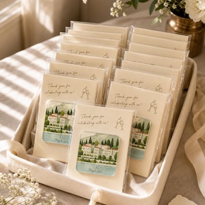 May include: A collection of rectangular party favours in clear plastic sleeves, each featuring a cream-coloured card with the text "Thank you for celebrating with us!" and a graphic of two wine glasses, paired with a square magnet showing a villa.