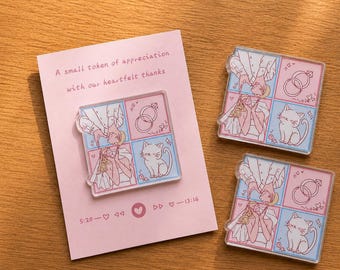 May include: Three clear acrylic pins with a pink and blue grid design featuring a couple, rings, and a cat. A pink card with text that reads "A small token of appreciation with our heartfelt thanks" is also present.