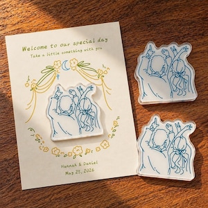 May include: A wedding invitation card with the text "Welcome to our special day" and "Take a little something with you". The card is decorated with floral and moon illustrations. Three clear acrylic wedding favors with a blue line drawing of a couple are also present.
