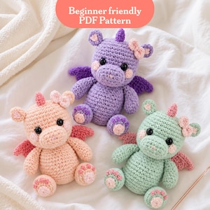 May include: Three crocheted dragon toys in pastel colors: peach, purple, and mint green. Each dragon has wings, a horn, and a bow. The text "Beginner friendly PDF Pattern" is in a peach oval at the top. The toys are on a white, textured surface.