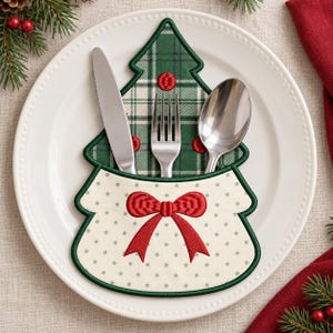 May include: A Christmas tree-shaped fabric cutlery holder featuring a green plaid pattern on the upper section and a cream-colored base with small green stars and a red embroidered bow, holding a silver knife, fork, and spoon.