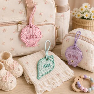 以下が含まれることがあります： ピンク、ミントグリーン、ラベンダー色の貝殻の形をした刺繍入りバッグタグ3点。EMMA、AVA、OLIVIAの名前入り。タグには水玉模様や花柄があしらわれ、マザーズバッグ、布、ポーチに取り付けられています。