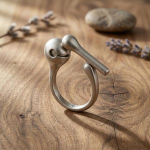 Puede incluir: Un anillo plateado con un diseño abstracto único. El anillo presenta una parte superior redondeada con una pequeña abertura, conectada a una banda curva. Un elemento cilíndrico recto se extiende desde la parte superior. El anillo se muestra sobre una superficie de madera.