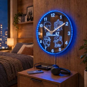 Puede incluir: Un reloj de pared circular con un borde de luz LED azul y una esfera negra con ilustraciones de líneas blancas de cuatro personajes. El centro muestra el texto GORILLAZ en blanco, con números blancos del 1 al 12 y manecillas iluminadas.