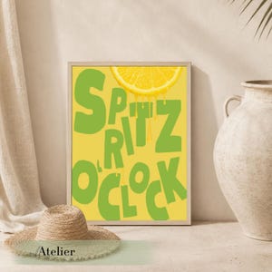 Puede incluir: Una impresión artística enmarcada con las palabras "Spritz O'Clock" en verde, con una rodaja de limón goteando jugo. El fondo es amarillo. Un sombrero de paja está en primer plano. Un jarrón beige está a la derecha.
