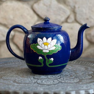 Könnte beinhalten: Eine blaue Teekanne mit einem weißen Seerosen-Design und goldenen Akzenten. Die Teekanne steht auf einem dekorativen Tablett.