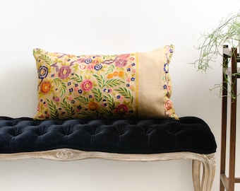 May include: A decorative pillow with a floral pattern in shades of pink, yellow, blue, and green on a cream background. The pillow has a gold trim and is sitting on a black velvet bench.