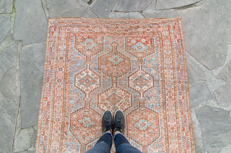 Puede incluir: Una alfombra vintage con un patr&oacute;n geom&eacute;trico en tonos rojos, azules y blancos. La alfombra tiene un aspecto desgastado y descolorido, y los colores son apagados. La alfombra est&aacute; colocada sobre una superficie de piedra.