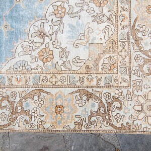 Op de afbeelding: Een vintage tapijt met een lichtblauwe achtergrond en ingewikkelde bloemmotieven in tinten bruin, beige en cr&egrave;me. Het tapijt heeft een rand met een repeterend geometrisch ontwerp.