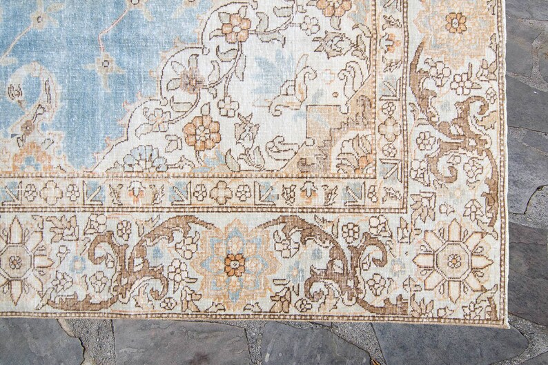 Op de afbeelding: Een vintage tapijt met een lichtblauwe achtergrond en ingewikkelde bloemmotieven in tinten bruin, beige en cr&egrave;me. Het tapijt heeft een rand met een repeterend geometrisch ontwerp.