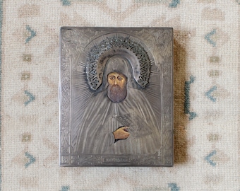 May include: A silver-toned icon with an image of a bearded man in a robe. The icon has an ornate border and a halo around the man's head. The text "Св. НАУМ ОХРИДСКИЙ" is written at the bottom of the icon.