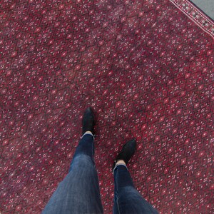 May include: A red rug with a geometric pattern and a person's legs wearing blue jeans and black shoes standing on the rug.