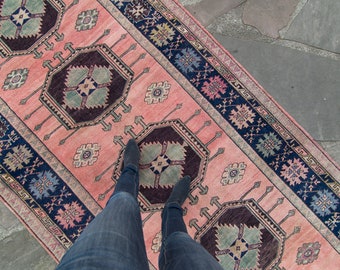 May include: A rectangular, vintage-style rug with a pink central field and dark blue borders. The rug features geometric patterns in shades of burgundy, teal, and pink. The rug is displayed on a stone surface.