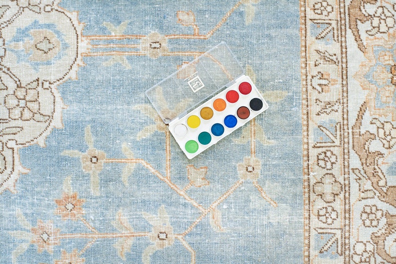 Op de afbeelding: Een plastic aquarelverfset met 12 kleuren, waaronder rood, oranje, geel, groen, blauw, paars en wit. Het verfset is open en ligt op een lichtblauw tapijt met een bloemenpatroon.