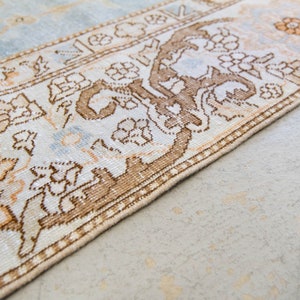 Op de afbeelding: Een lichtblauw en beige kleed met een bruin bloemenpatroon. Het kleed heeft een franje rand.