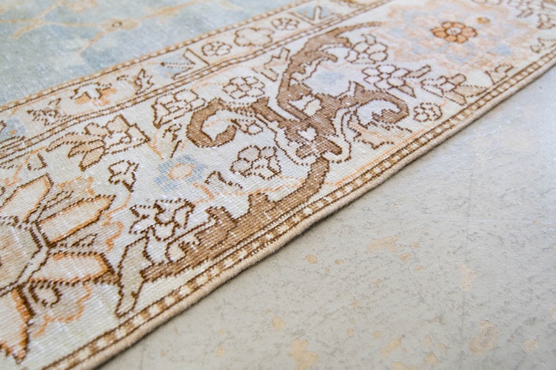 Op de afbeelding: Een lichtblauw en beige kleed met een bruin bloemenpatroon. Het kleed heeft een franje rand.