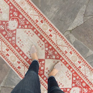 Może przedstawiać: Stopy osoby w beżowych butach z ozdobnymi szwami stoją na czerwono-kremowym dywaniku. Dywanik leży na szarej kamiennej podłodze.