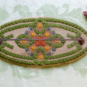 May include: An oval-shaped, beige fabric towel rack with a green embroidered design. The rack has a metal bar across the center and two metal knobs on either side.