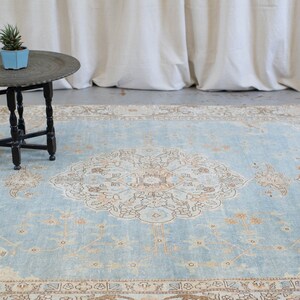 Op de afbeelding: Een lichtblauw tapijt met een beige en bruin bloemenpatroon. Het tapijt ligt op een betonnen vloer voor een wit gordijn.