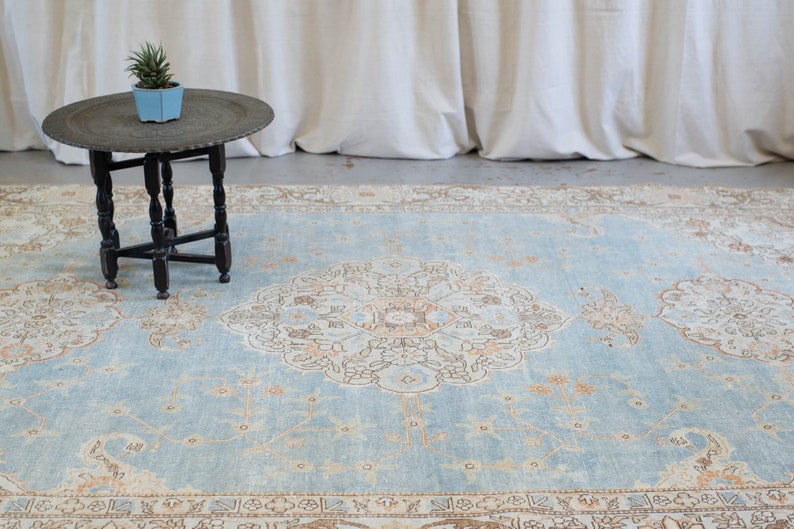 Op de afbeelding: Een lichtblauw tapijt met een beige en bruin bloemenpatroon. Het tapijt ligt op een betonnen vloer voor een wit gordijn.