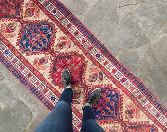 Puede incluir: Una alfombra de pasillo beige con un patrón geométrico rojo, azul y blanco. La alfombra está colocada sobre un suelo de piedra y las piernas de una persona están de pie sobre ella, con pantalones vaqueros azules y zapatos negros.
