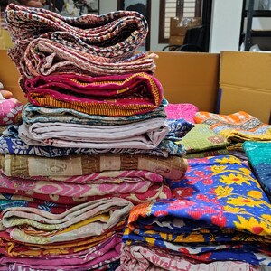 May include: A large stack of folded cotton quilts featuring various colorful patterns, including floral, geometric, and abstract designs in shades of pink, blue, yellow, and brown. Each quilt is stitched with visible running thread patterns.