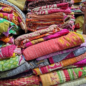 May include: A large stack of folded kantha quilts featuring a variety of vibrant colors, intricate patterns, and visible running stitch details. The textiles display diverse floral, geometric, and abstract prints in shades of pink, yellow, green, and blue.