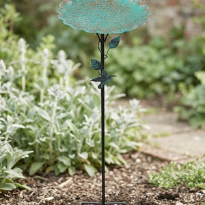 May include: A teal and bronze-colored bird bath on a black metal stand. The bird bath has a scalloped edge and a textured surface. A hummingbird and leaves are attached to the stand. The bird bath is in a garden setting.