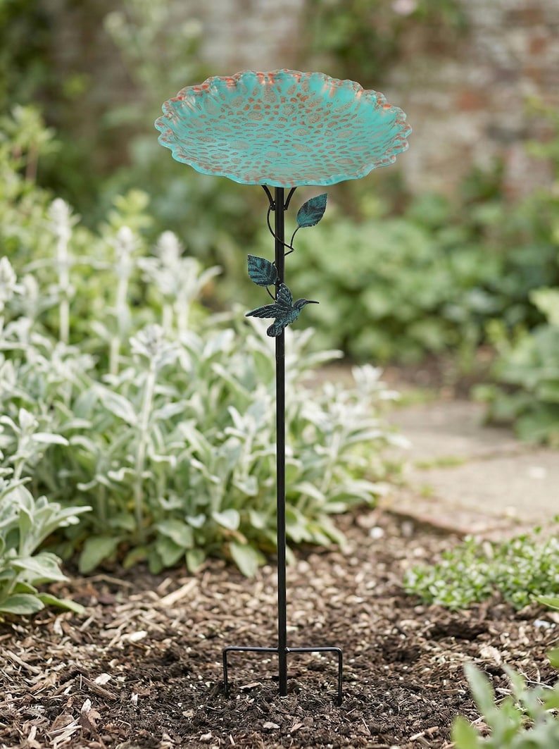 May include: A teal and bronze-colored bird bath on a black metal stand. The bird bath has a scalloped edge and a textured surface. A hummingbird and leaves are attached to the stand. The bird bath is in a garden setting.
