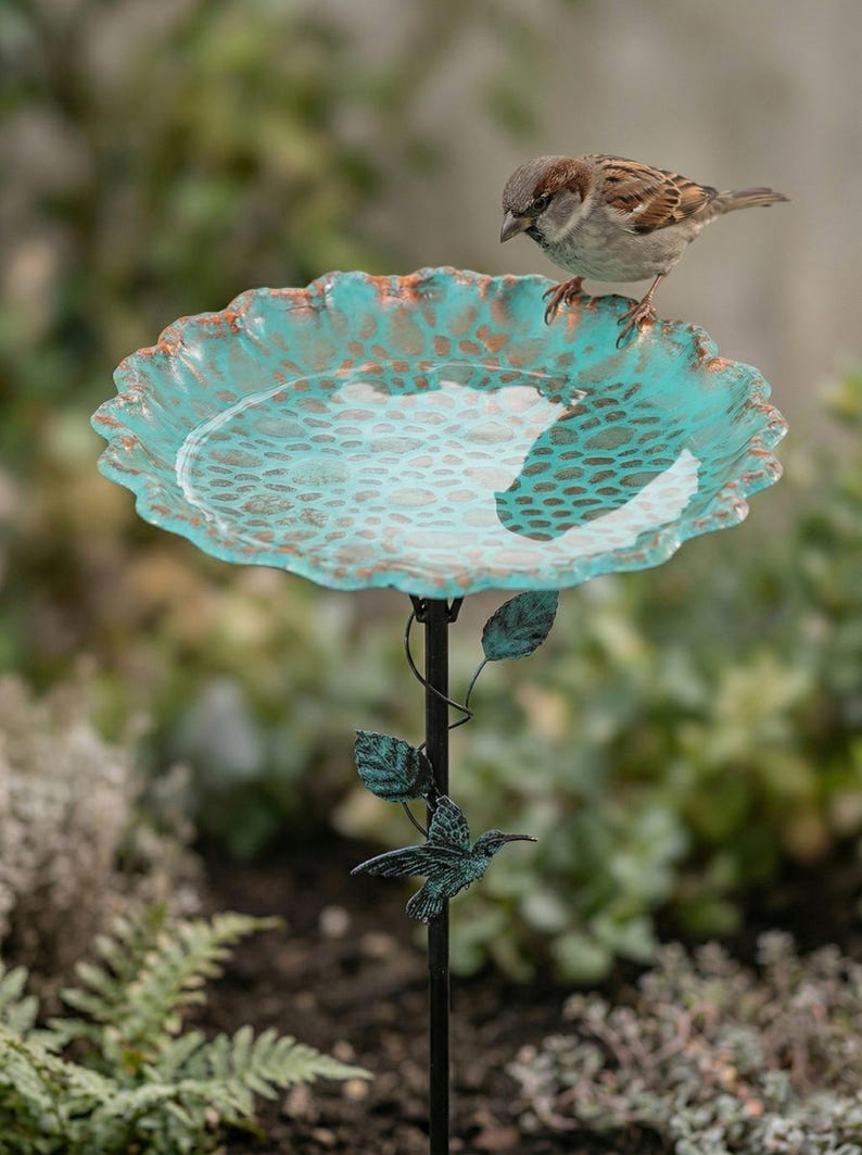 May include: A decorative bird bath with a turquoise and bronze-colored bowl, supported by a black metal stand. A small bird perches on the edge of the bowl. The stand features leaf and hummingbird accents, perfect for a garden.