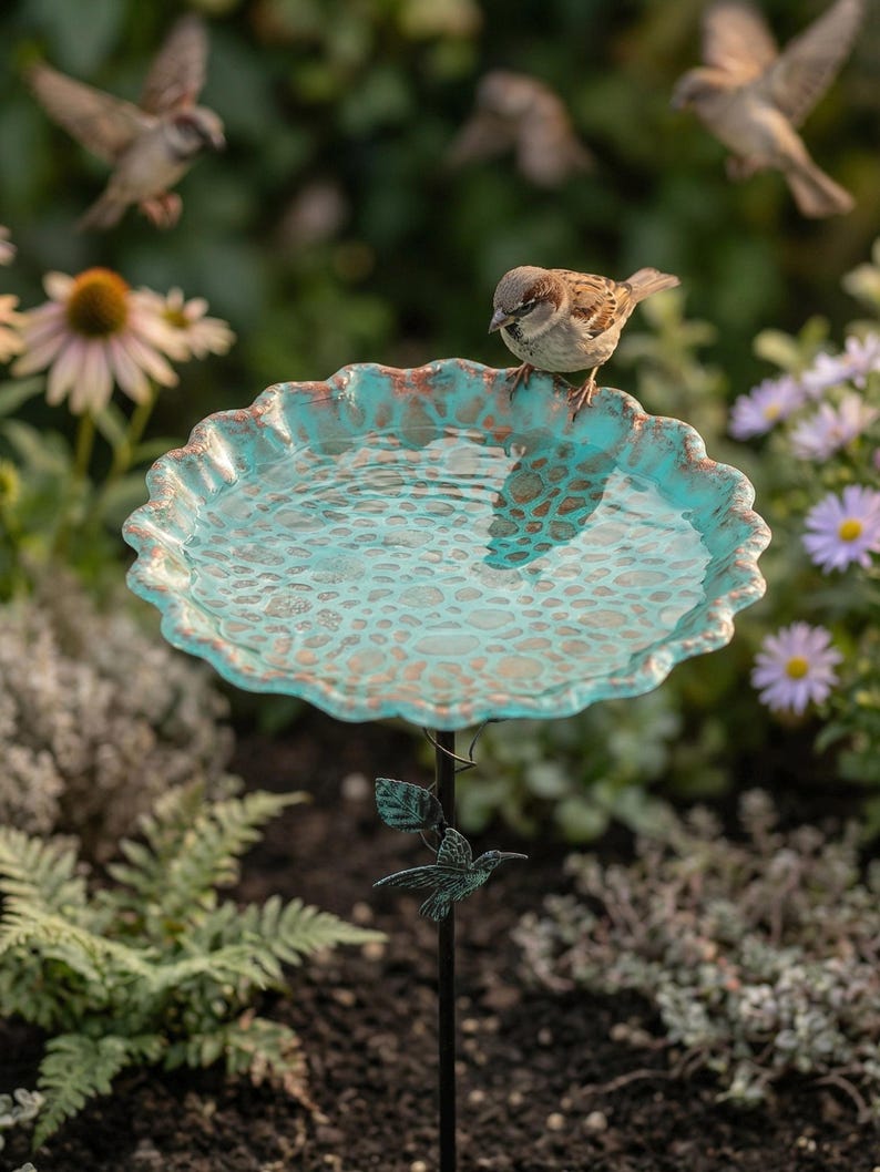May include: A decorative bird bath with a turquoise and bronze finish, featuring a scalloped edge. A small bird perches on the edge, with other birds flying in the background. The bird bath is supported by a black metal stand with a hummingbird accent.