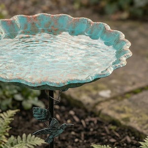 May include: A turquoise and copper-colored bird bath filled with water. The scalloped rim has a weathered appearance. A decorative hummingbird and leaf design adorns the black metal stand. The bird bath is set outdoors among greenery.