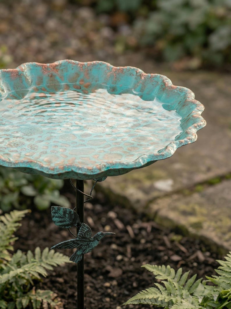 May include: A turquoise and copper-colored bird bath filled with water. The scalloped rim has a weathered appearance. A decorative hummingbird and leaf design adorns the black metal stand. The bird bath is set outdoors among greenery.