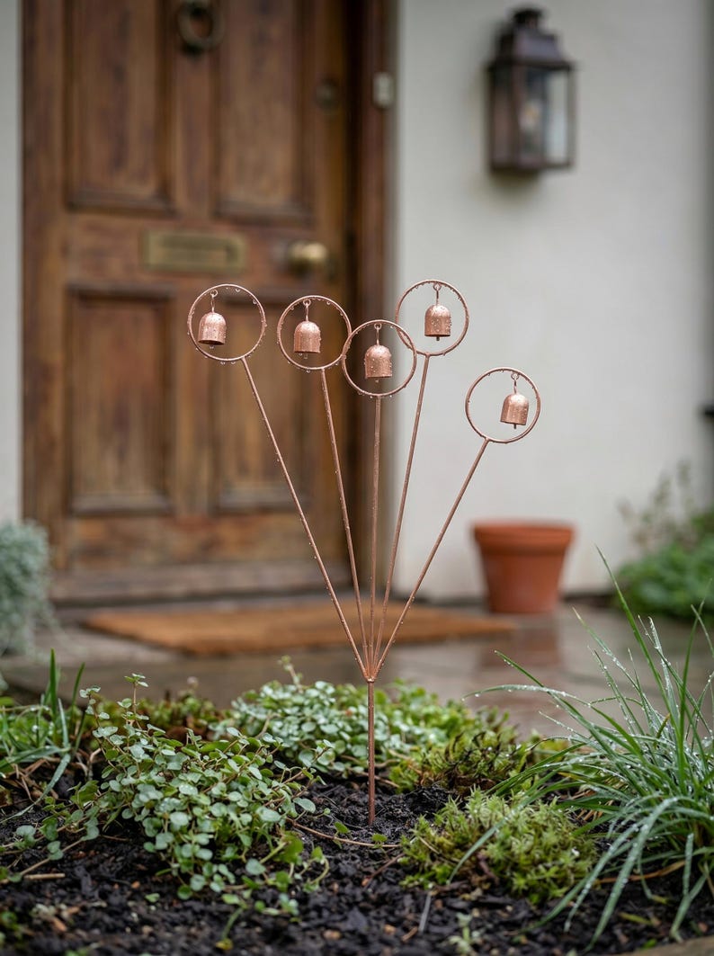 May include: Copper-colored garden stakes, each topped with a small bell inside a circular frame. The stakes are arranged in a fan shape, planted in a bed of green plants. The background includes a wooden door and a terracotta pot.