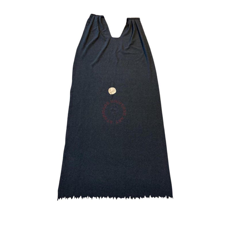 Op de afbeelding: Een donkergrijze cape met een rafelige zoom. De cape heeft een brede halslijn en een cirkelvormig embleem in het midden. De stof lijkt een zacht, draperend materiaal te zijn.