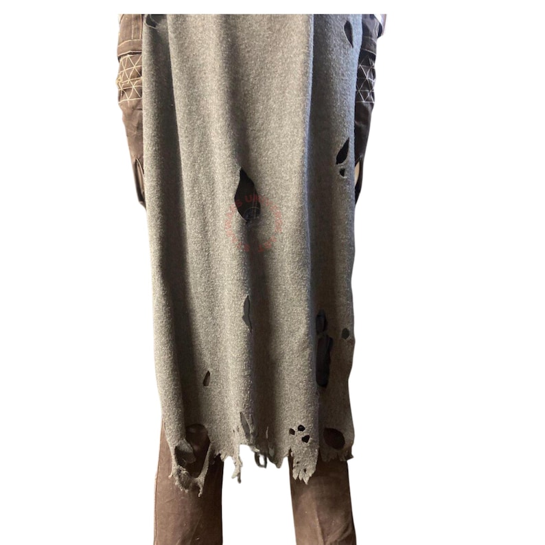 Op de afbeelding: Een grijs, versleten stoffen kledingstuk met meerdere gaten en een gerafelde onderrand. Het kledingstuk wordt gedragen over een bruine broek met decoratieve stiksels.