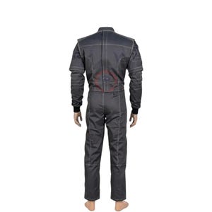 May include: A dark gray racing suit featuring long sleeves with black ribbed cuffs, a high collar, and prominent white contrast stitching throughout the garment. The back of the suit displays a circular red logo graphic centered on the upper back area.