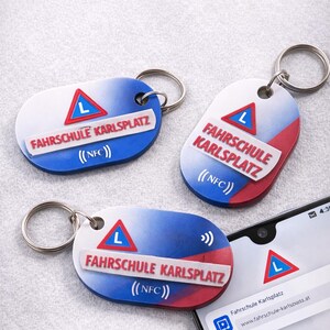 Op de afbeelding: Drie sleutelhangers met de tekst "FAHRSCHULE KARLSPLATZ" en een rood-blauw ontwerp. Elke sleutelhanger heeft een zilveren ring en de tekst "(NFC)". Een smartphone toont een melding met hetzelfde logo.