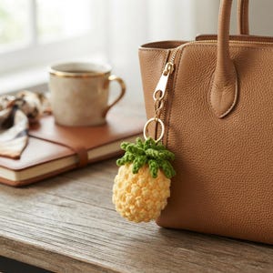 May include: A crochet pineapple keychain featuring a textured yellow body and green leafy top, attached to a gold-toned metal ring and clasp. The accessory is clipped onto the side of a tan leather handbag with a visible zipper and pebbled texture.