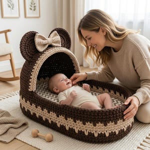 May include: A dark brown and beige crocheted baby bassinet with a hood shaped like rounded ears and a decorative bow. The bassinet features a chevron-patterned mattress pad and a thick, textured yarn construction with a two-tone color scheme.