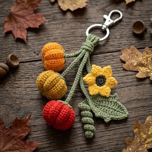 May include: A crochet keychain featuring three small pumpkins in orange, yellow, and red, alongside a yellow sunflower with a dark centre and a green leaf, all attached to a silver metal lobster clasp and ring on green cord stems.