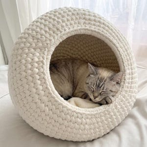 May include: A spherical, off-white crocheted pet bed with a large circular opening. Inside the textured, thick-yarn structure, a light-colored cat with grey tabby markings is curled up and sleeping peacefully on a soft, cream-colored cushion.