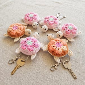 May include: A collection of five crocheted turtle keychains in shades of pink, orange, and cream. Each turtle features a floral-patterned shell, small black eyes, and a metal lobster clasp. Two of the keychains are attached to silver metal keys.
