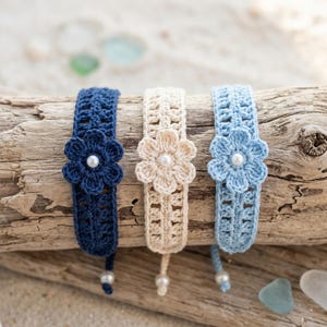 Può includere: Tre braccialetti all'uncinetto in blu navy, crema e azzurro. Ogni braccialetto presenta un motivo floreale centrale a sei petali con una piccola perla bianca al centro. Le fasce hanno un motivo traforato e una perla all'estremità.