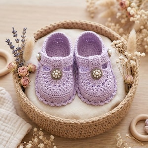 Peut inclure: Une paire de chaussures pour bébé en crochet violet clair avec une bride en T, des bords festonnés et un bouton décoratif central orné d'un groupe de petites perles blanches. Les chaussures sont présentées dans un panier tressé rond.