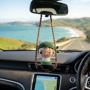 Könnte beinhalten: Gehäkelter Autoanhänger mit einer Figur mit grünen Haaren, grünem Hut und einer Narbe am Auge. Der Anhänger hängt an einem braunen Seil im Auto. Im Hintergrund ist eine Küstenstraße und das Meer zu sehen.