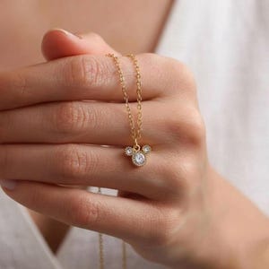 Puede incluir: Un collar dorado con un colgante con forma de cabeza de rat&oacute;n. El colgante presenta una gema grande, redonda y transparente en el centro, con gemas m&aacute;s peque&ntilde;as a cada lado. El collar se sostiene en una mano.