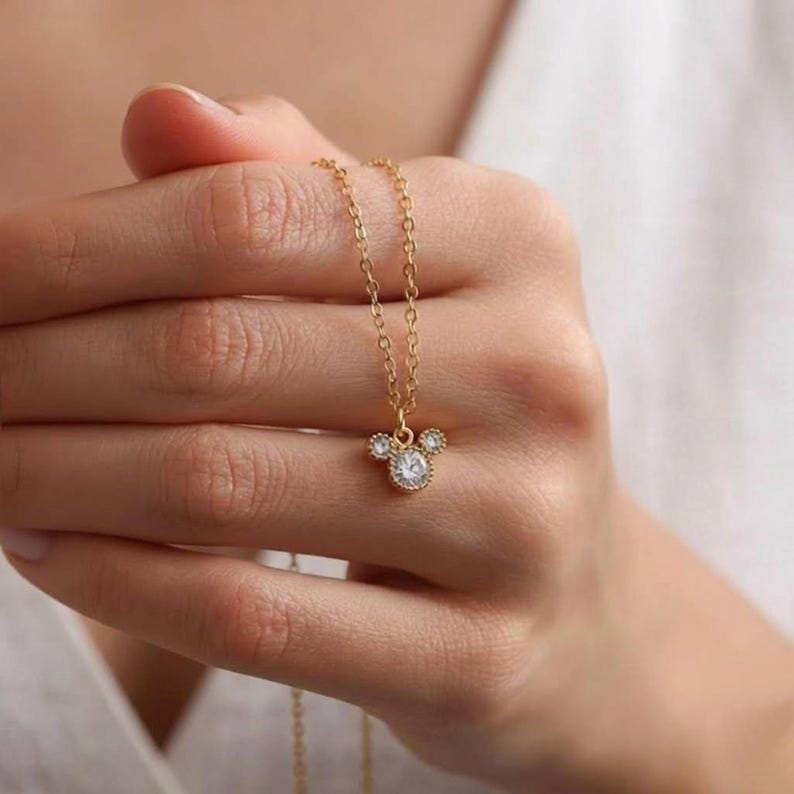 Puede incluir: Un collar dorado con un colgante con forma de cabeza de rat&oacute;n. El colgante presenta una gema grande, redonda y transparente en el centro, con gemas m&aacute;s peque&ntilde;as a cada lado. El collar se sostiene en una mano.